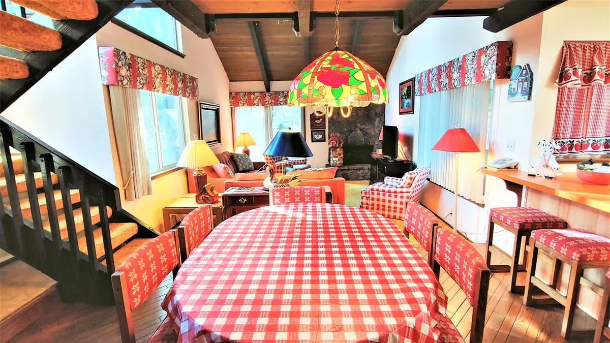 A wide shot features a checkered tablecloth dining table set beneath a stained glass lamp, with a comfortable seating area and a stone fireplace visible nearby. The view seamlessly blends dining and lounging spaces, reinforcing the open-concept layout.