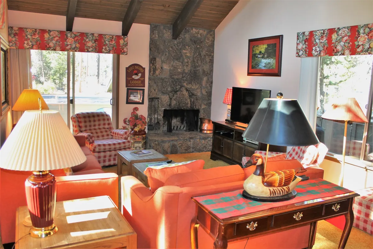 This photo spotlights the living room, centered around a classic stone fireplace with plaid sofas and a wooden coffee table. Decorative touches like a lamp and wall art complement the cozy, retro vibe, while large windows hint at an airy, sunlit atmosphere.