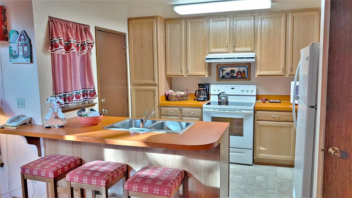 The image showcases a kitchen island with red checkered curtains and patterned bar stools, set against the backdrop of warm wooden cabinetry. The island provides a spot for casual snacks or socializing, tying in retro flair with practical amenities.