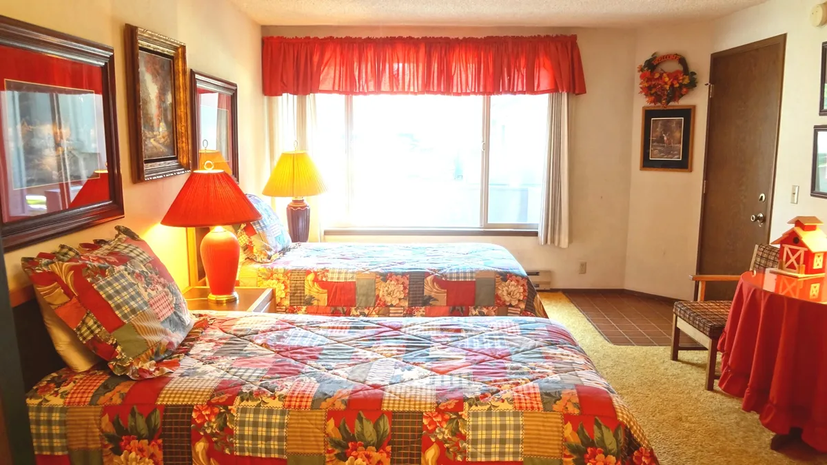 This image features two beds with colorful quilts in a sunlit bedroom, accompanied by a bedside table with a red lamp and decorative wall art. The large window allows plenty of natural light to flood the room, giving it a cozy and welcoming ambiance.