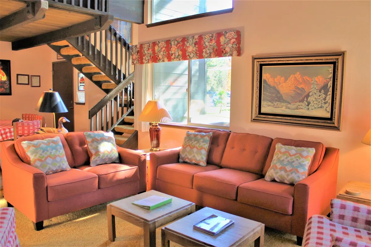 The image features a cozy living space with orange sofas, decorative cushions, a wooden staircase, framed painting, and a table lamp. Ample natural light fills the room, and the arrangement invites relaxation, conversation, or reading in a vintage atmosphere.