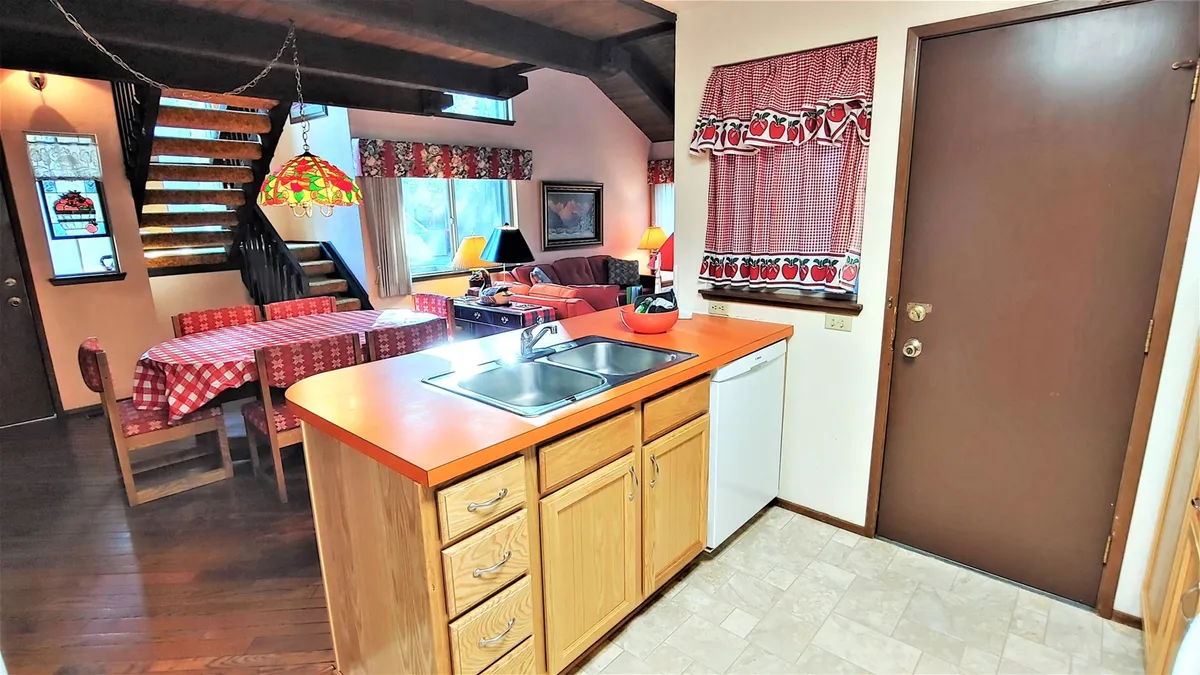 This interior photo captures the heart of the living space, featuring a welcoming kitchen counter, dining table, staircase, and a glimpse of the inviting living area. Warm lighting and retro touches set a nostalgic, homey atmosphere, making it easy to envision relaxing or gathering after a day of resort activities.