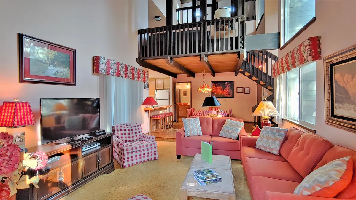 The image showcases a welcoming living room with a plush sofa, modern television, warm lighting, and a stylish staircase leading to the second level. The space feels open and inviting, perfect for relaxing, socializing, and unwinding after a day of Sunriver adventures.