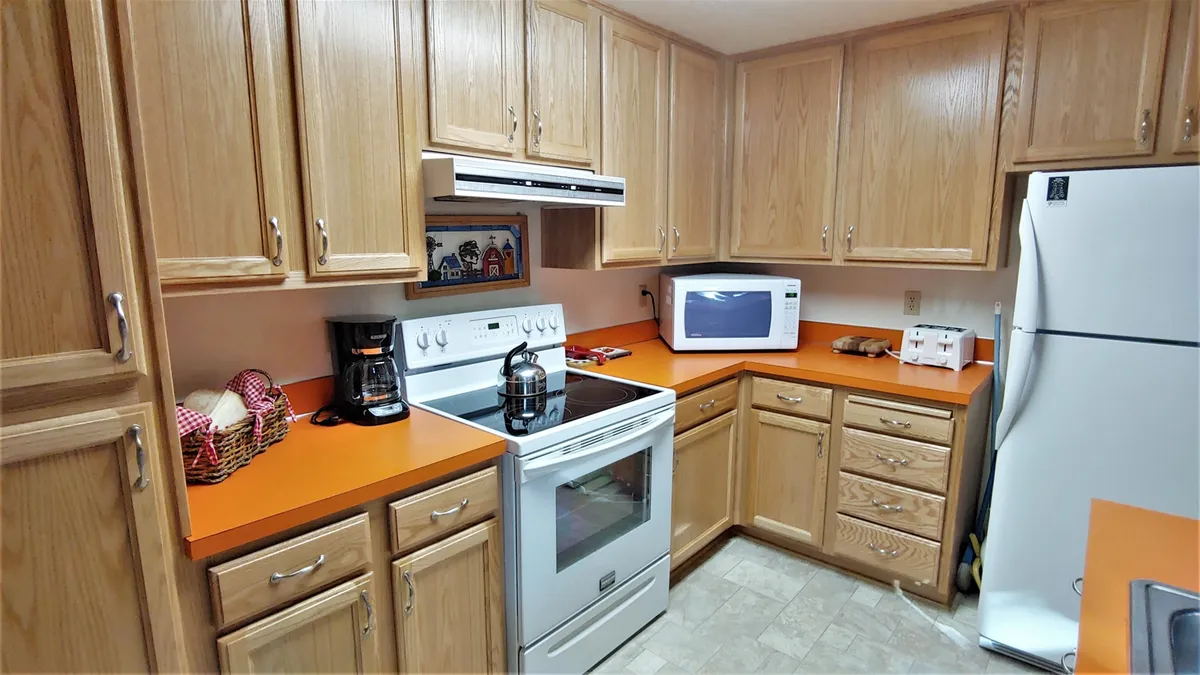 A bright, vintage kitchen with an orange countertop, white appliances, wooden cabinetry, and a coffee maker. The kitchen island suggests a space where family members gather to cook, bake, and enjoy morning coffee together.