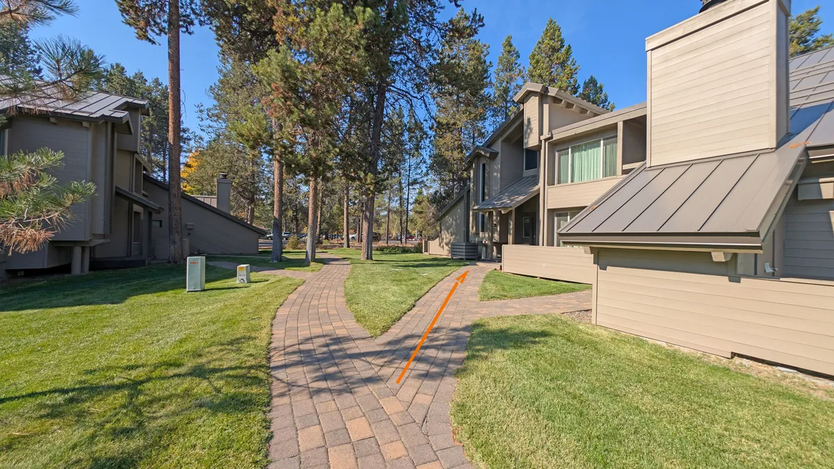 A tranquil exterior view of a pathway meandering through tall trees and between buildings. The setting is peaceful, with dappled sunlight and the sense of connection between nature and the community's outdoor amenities.