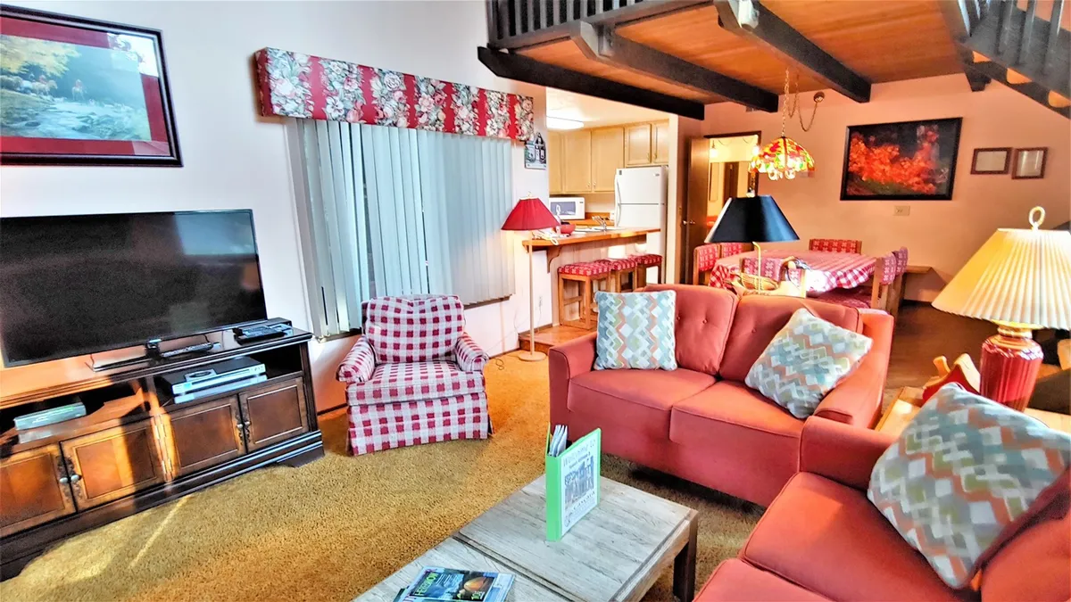 A comfortable living area with a red couch, checkered armchair, coffee table with books, TV, and a dining area in the background. The space is designed for lounging, reading, and spending time with family.