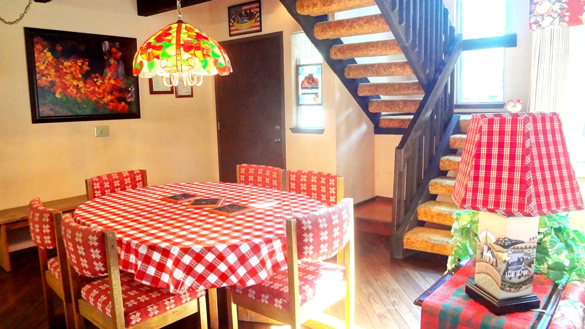 This image shows a welcoming dining area with a checkered tablecloth, a colorful lamp, a wooden staircase, and decorative artwork. The dining table is set in a cozy nook that feels perfect for family meals, game nights, or sharing stories with friends. The visible staircase hints at the unique two-level layout.