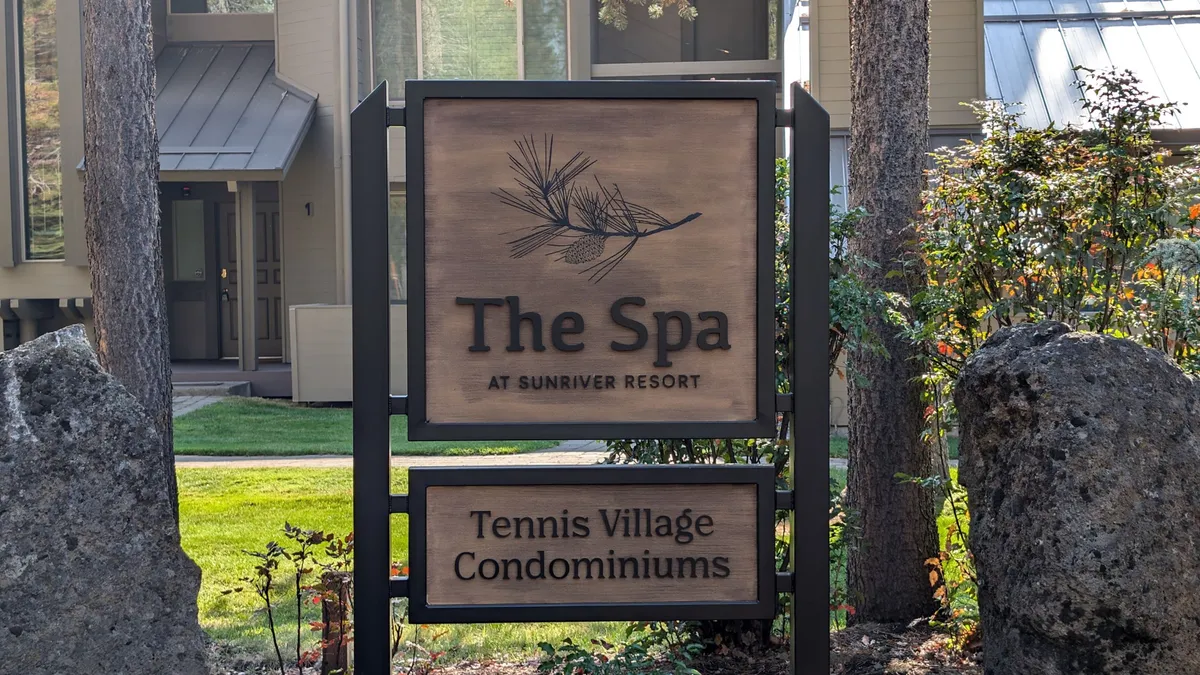 The exterior of the Sunriver resort spa, featuring a prominent 'The Spa' sign set amidst natural surroundings with rock features and green landscaping. Tennis courts are visible in the background, suggesting a variety of recreational activities available to guests.