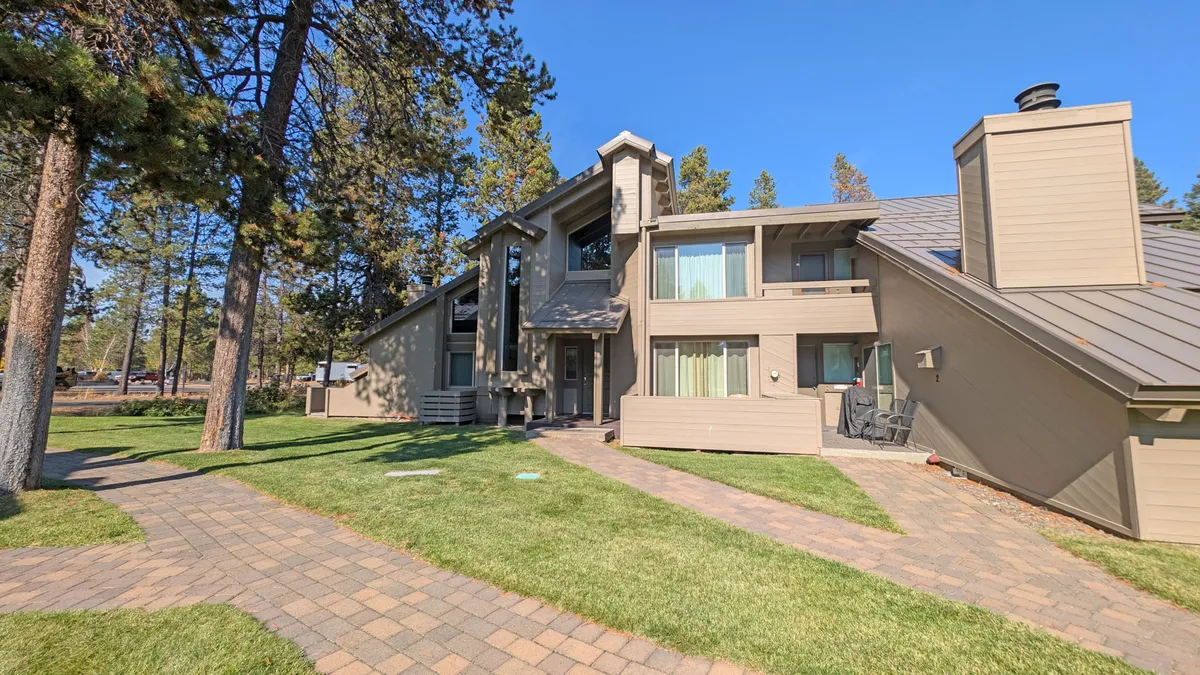 This image features the modern architecture of the condo building, highlighting the pathway leading to the entrance and the surrounding tall trees. The greenery and expansive sky suggest a natural, inviting environment and easy access to outdoor spaces.